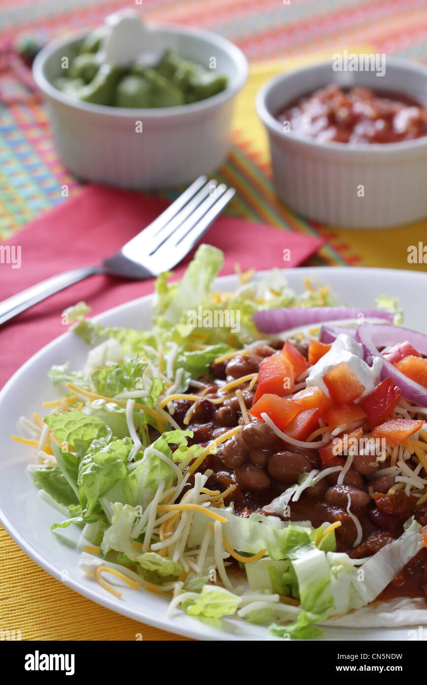 Ein Chili Bohnen Tostada mit Tomaten und saure Sahne auf einem Bett aus knackigen Salat mit Guacamole und heiße Soße Begleitungen Stockfoto