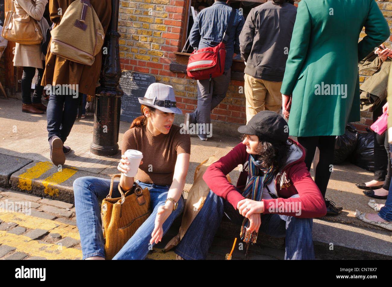 Zwei Mode-bewusste junge Frauen sitzen auf dem Bürgersteig in der Columbia Road, East London im Gespräch Stockfoto