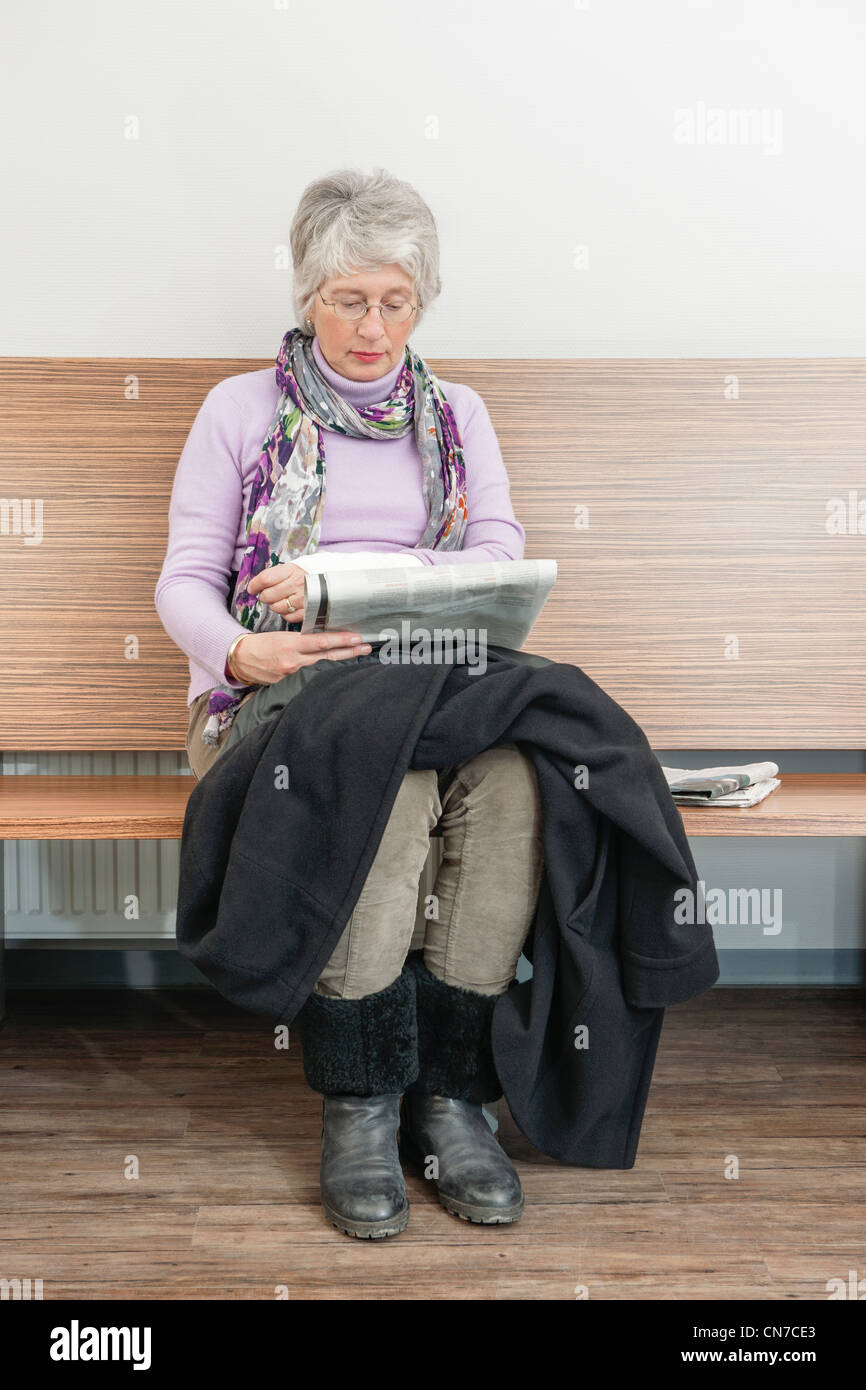 Frau liest eine Zeitung, sitzen auf einer Bank in einem Wartezimmer mit ihren Mantel auf ihrem Schoß und Beine gekreuzt. Stockfoto