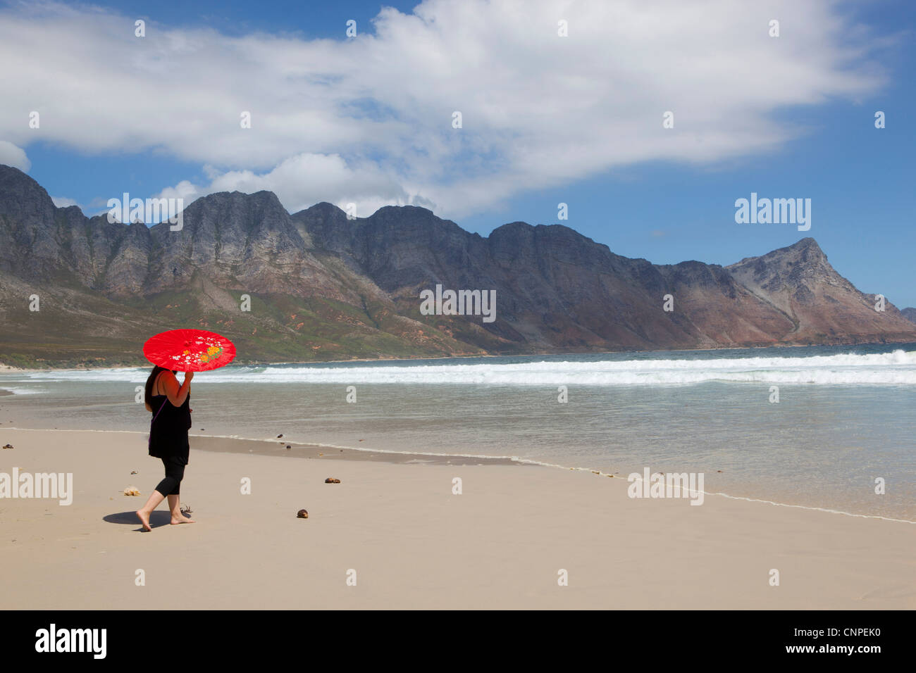 Eine Frau mit einem roten Sonnenschirm zu Fuß am Strand von Kogel Bay, Western Cape, Südafrika Stockfoto