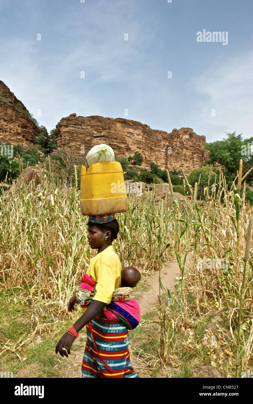 Eine Frau gleicht Gegenstände auf dem Kopf während des Gehens nach Hause mit ihrem Baby in Dogon County, Mali. Stockfoto