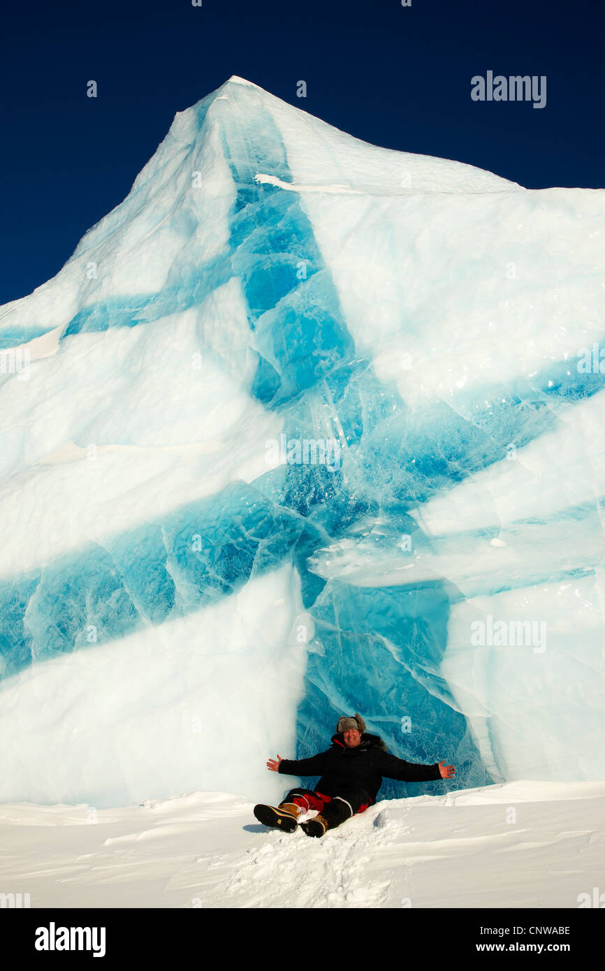 lächelnder Mann mit den Armen verteilt sitzen im Schnee vor Eisberg mit blauen Adern, Grönland, Ostgroenland, Tunu, Kalaallit Nunaat, Liverpool Land Lillefjord Stockfoto
