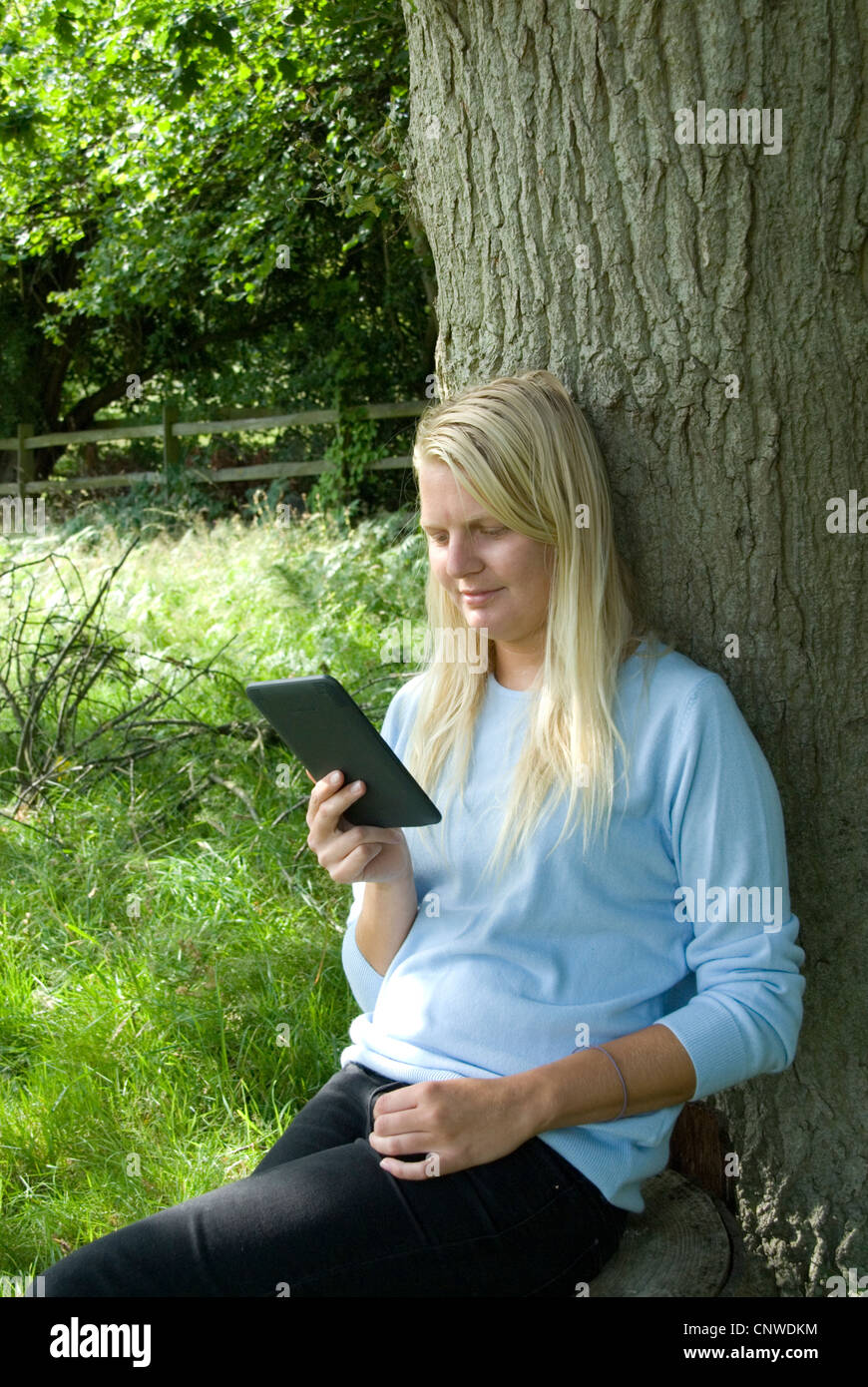 Blonde Mädchen liest einen Amazon Kindle elektronische e-Reader im Freien an einen Baum Hayley Williamson Herr gelehnt Stockfoto
