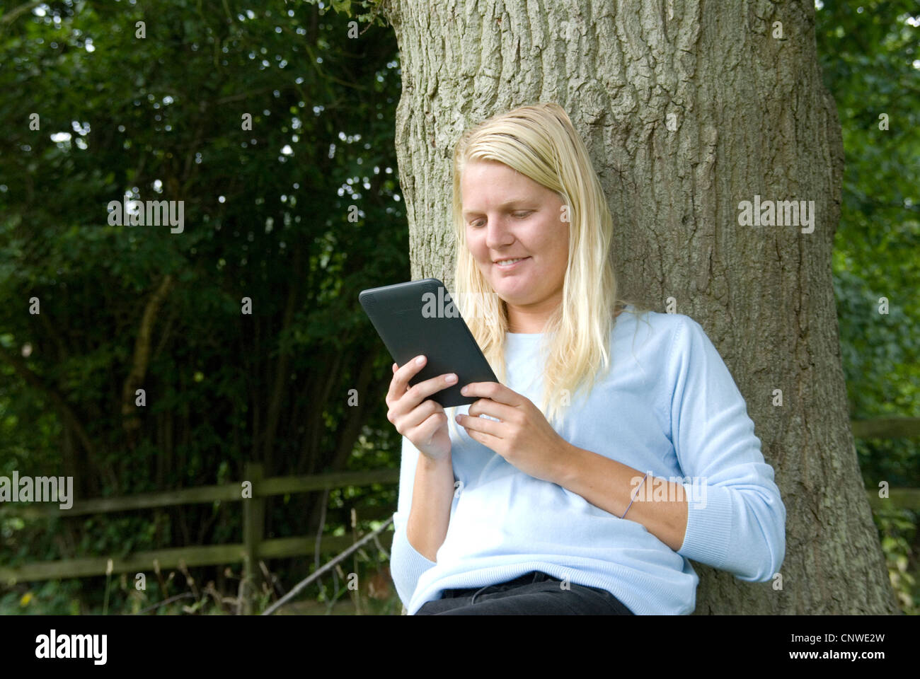 Blonde Mädchen liest einen Amazon Kindle elektronische e-Reader im Freien an einen Baum Hayley Williamson Herr gelehnt Stockfoto