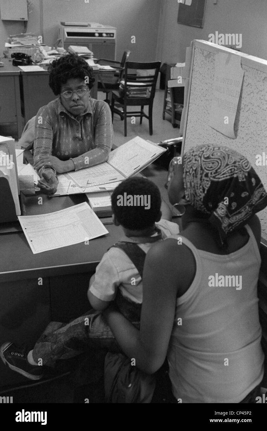 Eine afrikanische amerikanische Frau mit einem Kind auf dem Schoß im Gespräch mit einem Sachbearbeiter beim Sozialamt ein New York City. 14. Mai 1976. Stockfoto