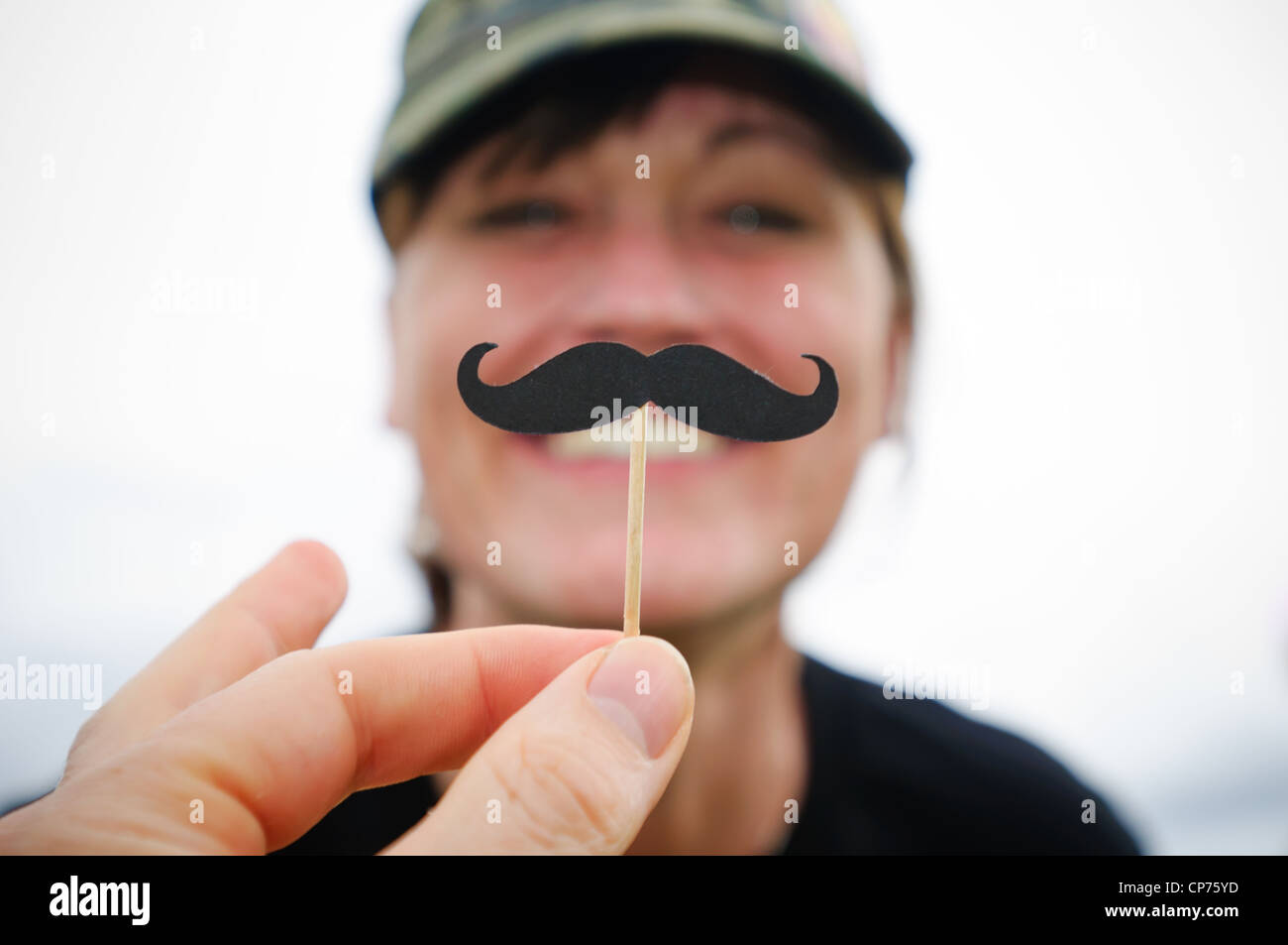 Eine Hand hält einen falschen Schnurrbart vor einer lächelnden Frau. Stockfoto