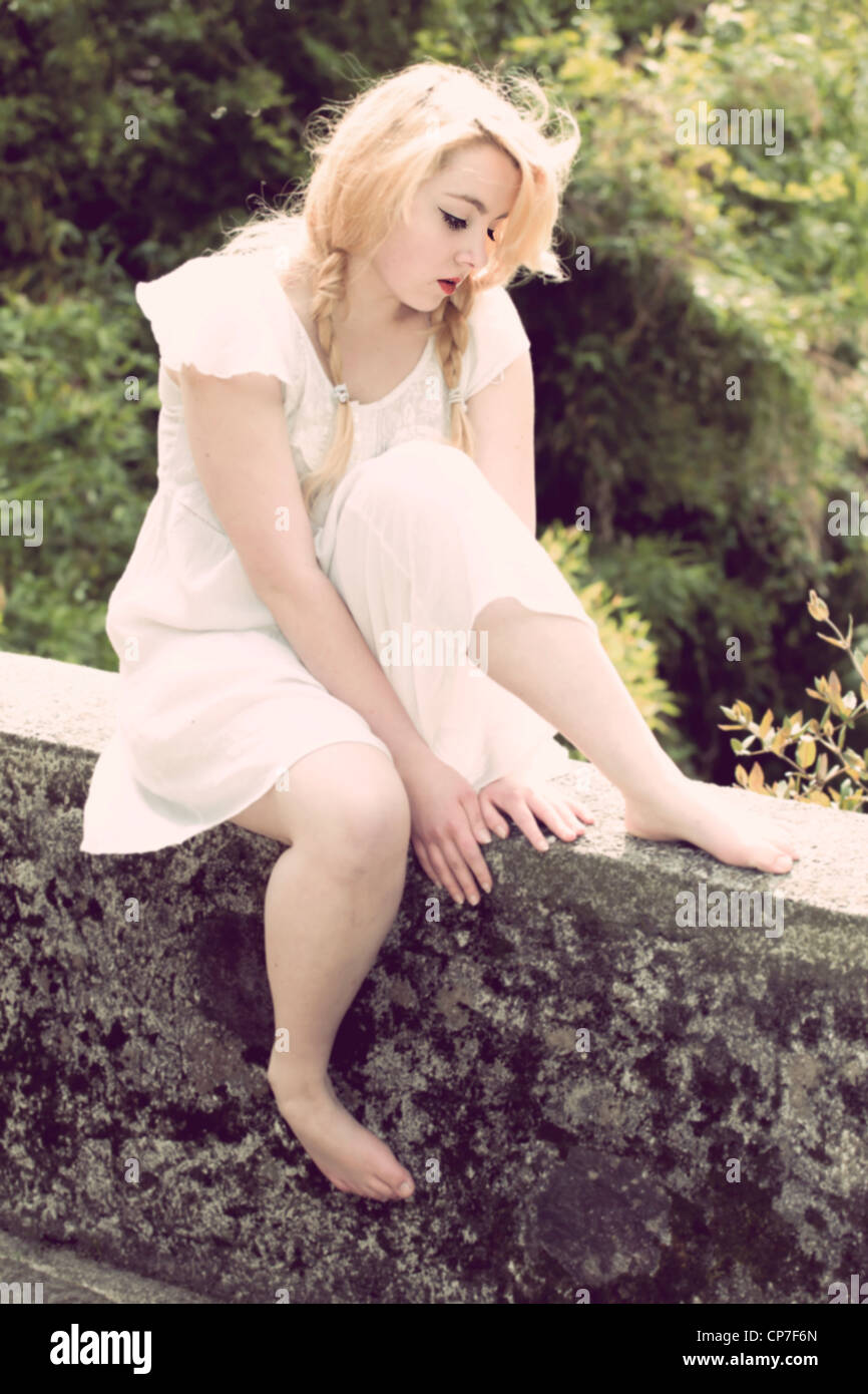 junge Frau mit Zöpfen sitzt auf einer Wand in einem weißen Kleid Stockfoto