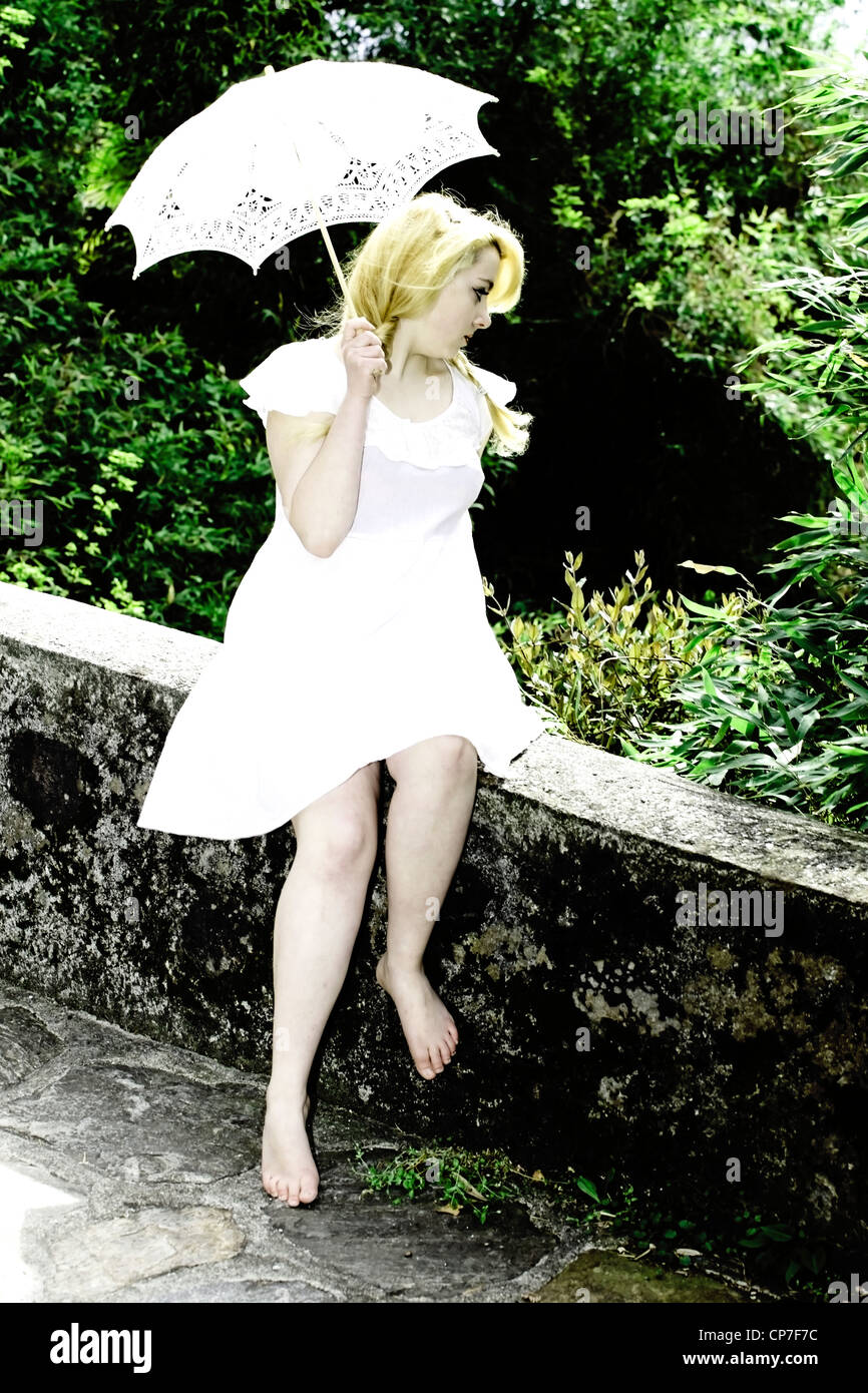 junge Frau im weißen Kleid sitzt auf einer Mauer mit einem Sonnenschirm Stockfoto