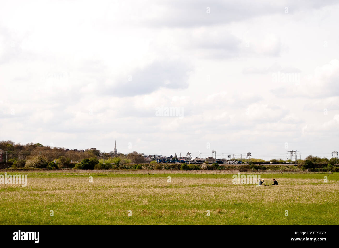 Zwei Menschen sitzen in einem Feld in Leyton Marsh mit der Industrielandschaft im Hintergrund, London, UK Stockfoto