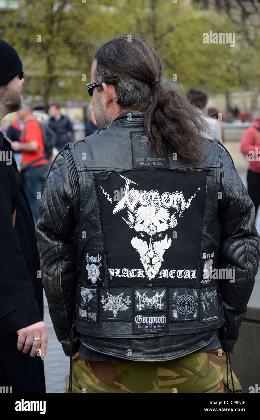 Mann mittleren Alters mit Pferdeschwanz trägt eine Leder Jacke Werbung Black Metal zweite Album Venom. Stockfoto