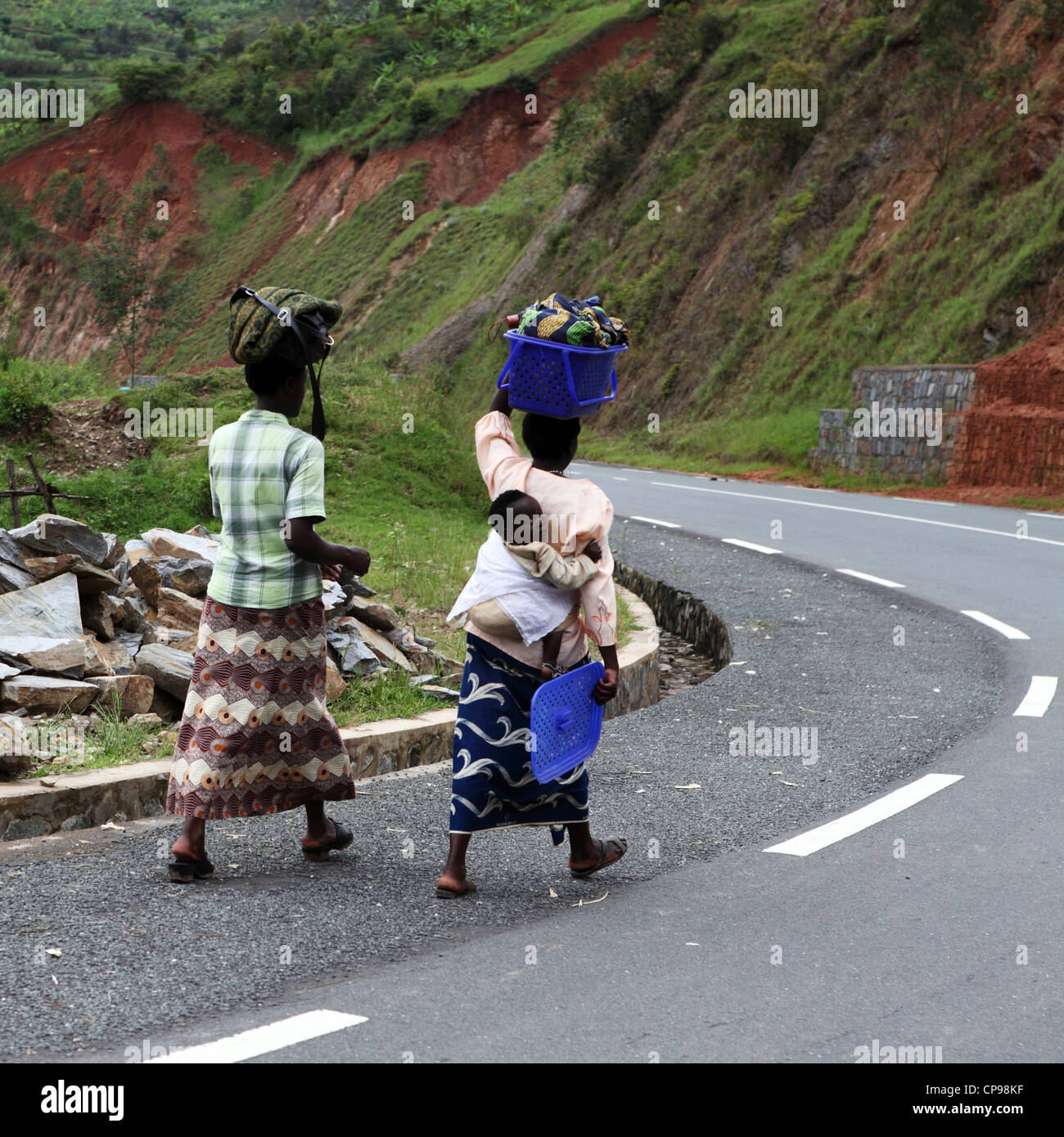 Frauen tragen waren auf den Kopf und man trägt ein Baby auf dem Rücken im ländlichen Ruanda. Die Frauen gehen an einem Straßenrand. Stockfoto