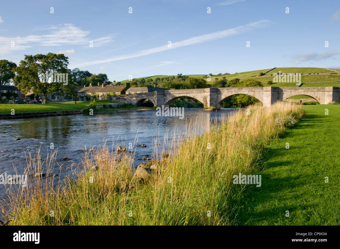 Steinerne Brücke über dem Fluss Wharfe, wie es fließt durch Burnsall Dorf- & malerische Landschaft auf sonnigen Sommerabend - Yorkshire Dales, England, UK. Stockfoto