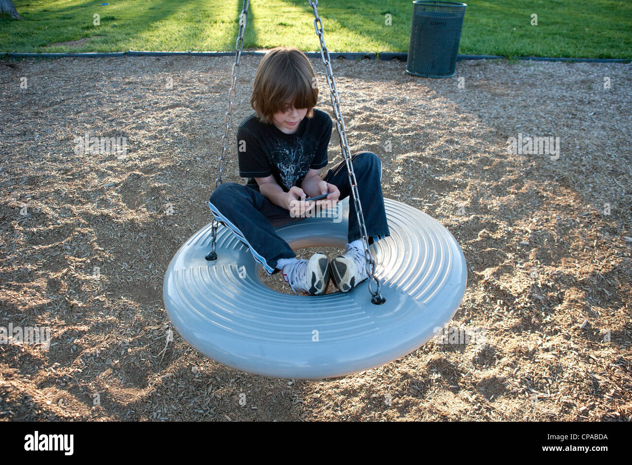 Neun Jahre altes Kind ein Spiel auf seinem Iphone beim Sitzen auf einem Donut-Schaukel auf einem Spielplatz. Stockfoto
