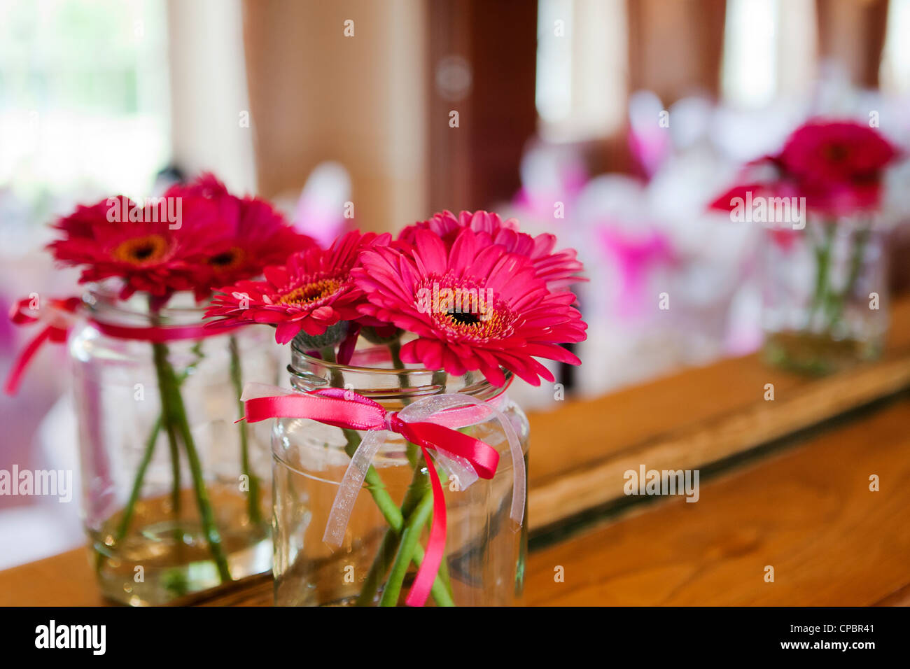 Rosa Gerberas in einer kleinen Vase, ein Glas Konfitüre, gebunden mit rosa Band in einem Spiegel bei einer Hochzeit reflektiert. Stockfoto
