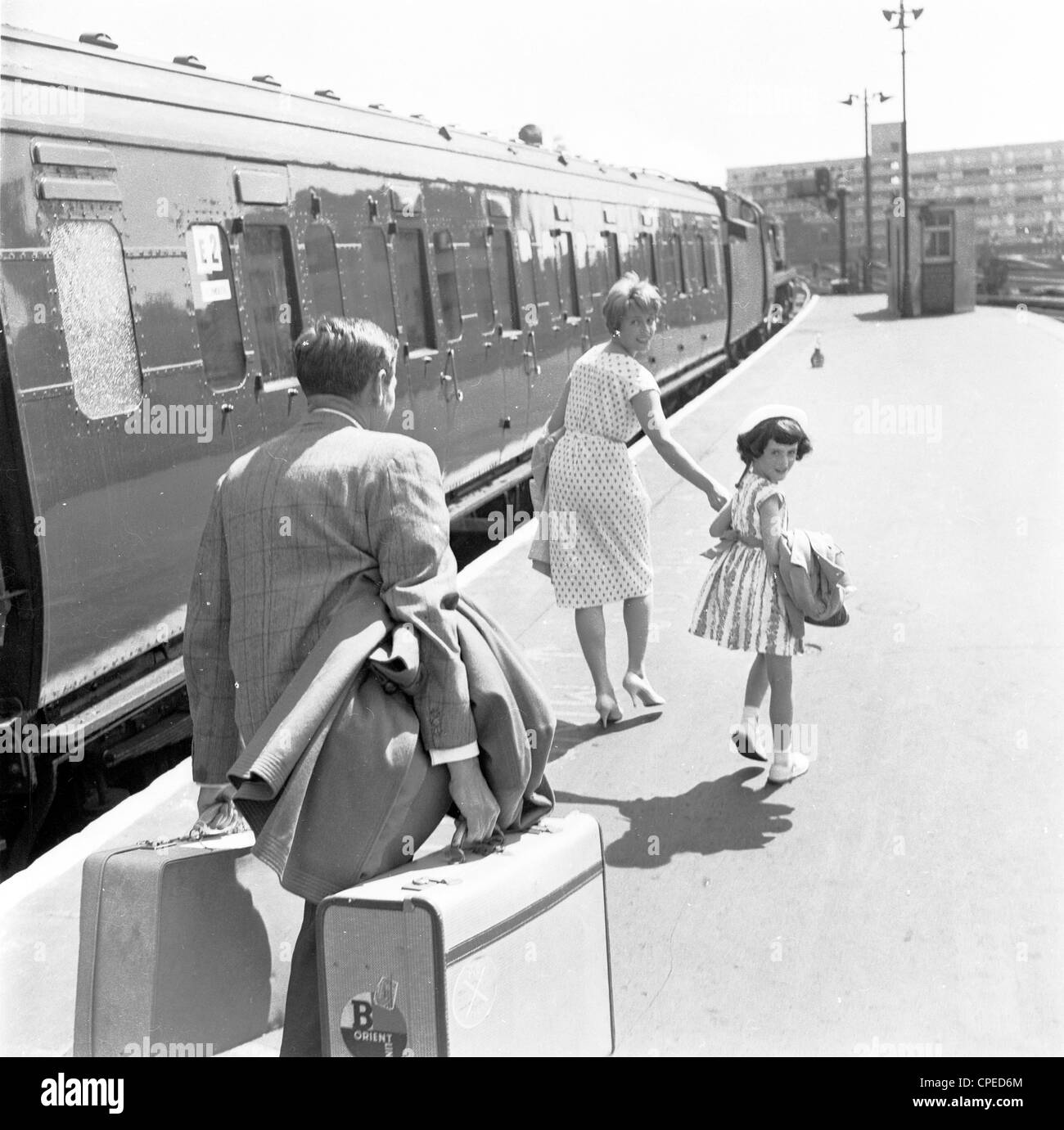 1960s, historisch, draußen auf einem leeren Bahnsteig, eine Familie eilt, um einen wartenden Zug zu cachieren, mit der Dame und der jungen Tochter vorne und mit dem passenden Ehemann hinten, der die Mäntel und das Gepäck trägt, zwei schwere Koffer in Boxenrahmen der Ära. Stockfoto