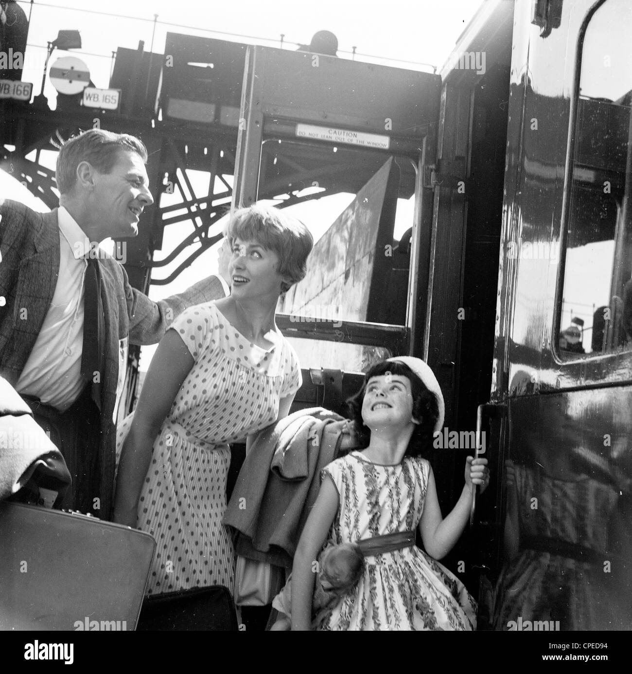 1960s, historisch, Sommerzeit eine aufgeregte Familie, die in den Urlaub fährt, an Bord eines Zugwagens auf einer Bahnplattform, England, Großbritannien. Die Dame trägt ein gepunktetes Kleid, das in dieser Zeit in Mode war, während der Mann formallt in Hemd und Krawatte gekleidet ist, mit Jacke, wie es damals üblich war. Die junge Tochter blickt aufgeregt zu ihren Eltern. Stockfoto