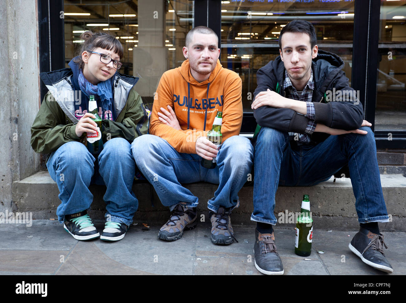 Tägliches Stadtleben: Drei junge Menschen sitzen auf einem Bürgersteig und trinken Bier auf der Brick Lane, London, England, Großbritannien. Stockfoto