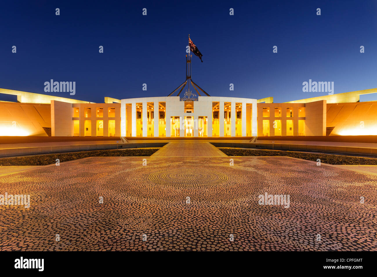 Parliament House, Canberra, Australien, in der Dämmerung beleuchtet. Aborigine-Mosaik im Vordergrund, ist australische Flagge. Stockfoto