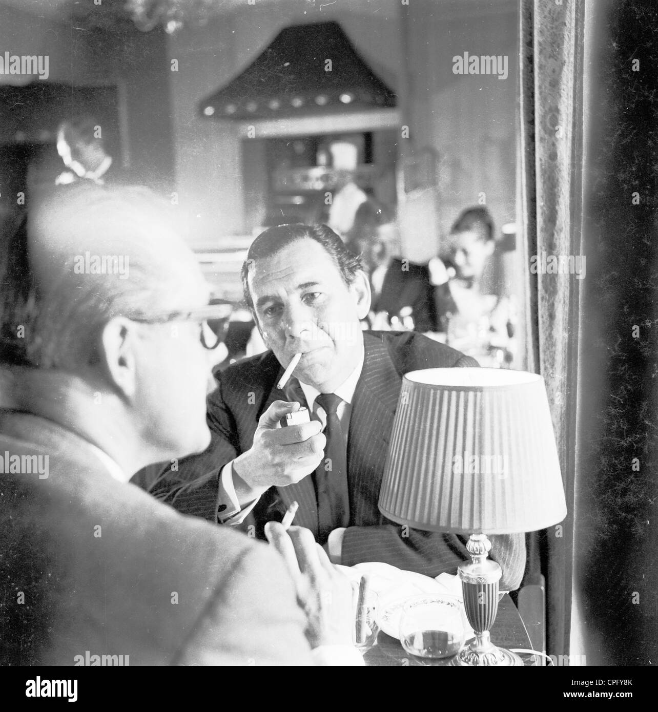 London, 1960er Jahre. Die Freuden des Rauchens. Ein Geschäftsmann mit einer Mahlzeit mit einem Kollegen in einem Restaurant leuchtet seine Zigarette. Stockfoto