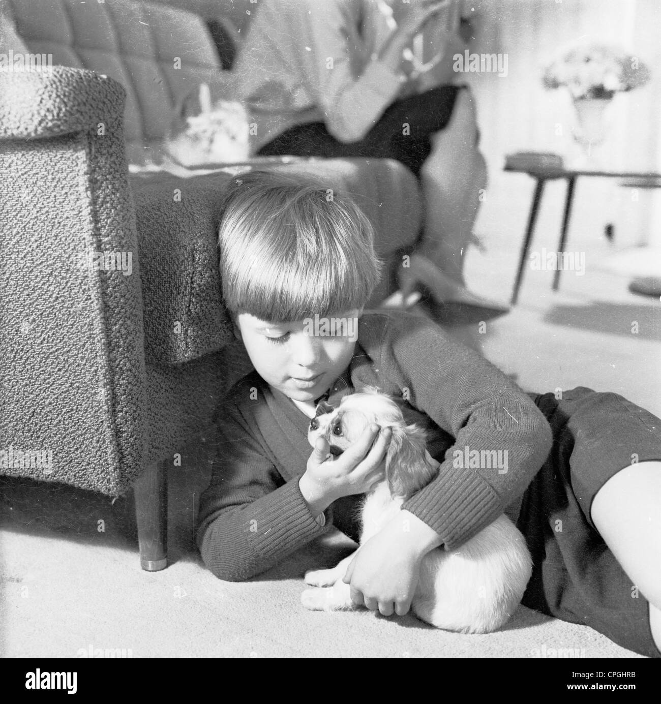 1960s, historisch, nach der Schule, ein kleiner Junge, der auf einem Wohnzimmerboden liegt und liebevoll seinen kleinen Welpen hält, während seine Mutter auf dem Sofa sitzt, England, Großbritannien. Stockfoto