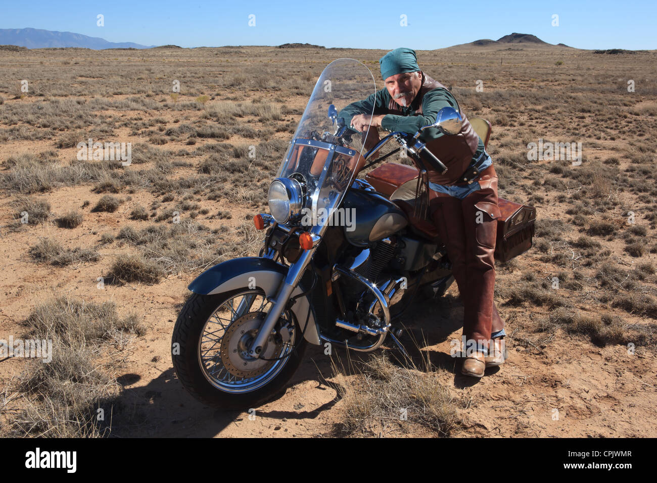 Ältere kaukasischen Mann mit braunem Lederchaps und Weste, beugte sich über den Lenker von seinem Motorrad in die offene Wüste. Stockfoto