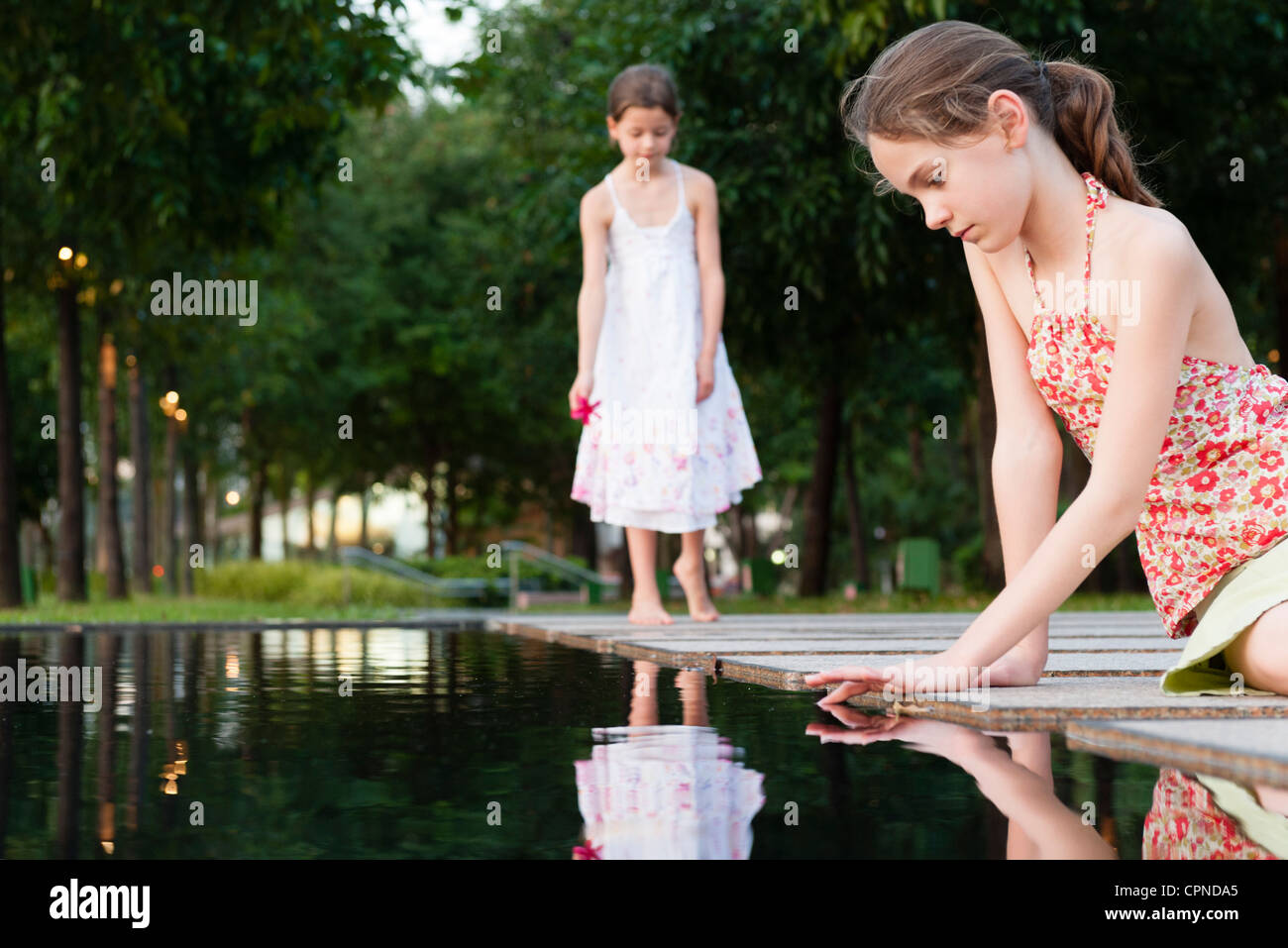 Mädchen sitzen durch Berühren der Oberfläche des Wassers, ein anderes Mädchen im Hintergrund Teich Stockfoto