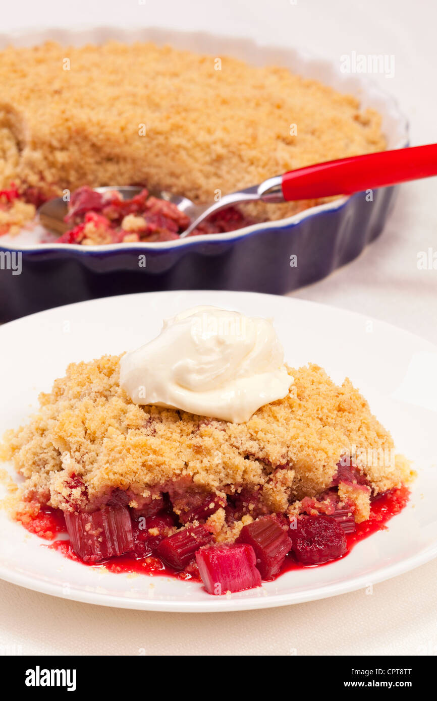 Ein Teller mit Rhabarber und Himbeeren crumble mit einem Klecks Sahne servieren Teller und Löffel im Hintergrund. Stockfoto