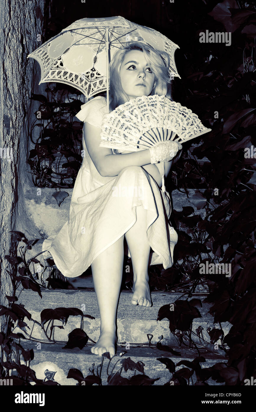 junge Frau sitzt auf einer Treppe mit Efeu und ein Sonnenschirm und ein Ventilator in ihren Händen hält Stockfoto