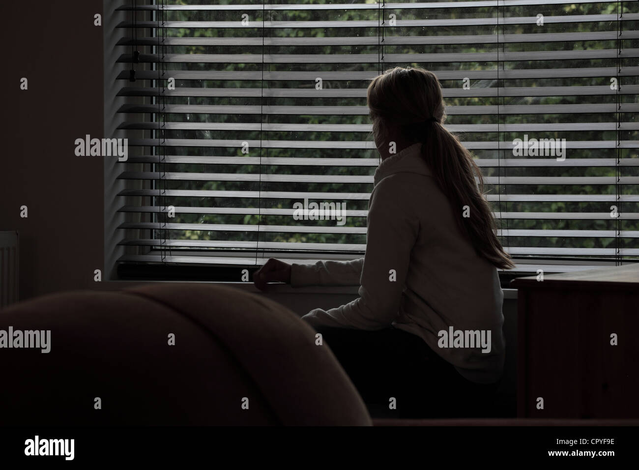 Junge Frau sitzt allein in einem dunklen Raum mit Blick durch ein Fenster Blind. Stockfoto