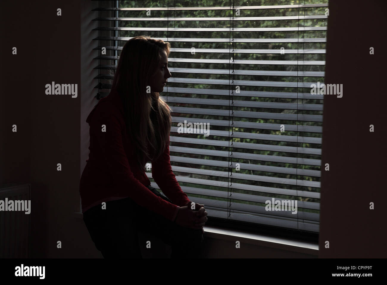 Junge Frau sitzt allein in einem dunklen Raum mit Blick durch ein Fenster Blind. Stockfoto