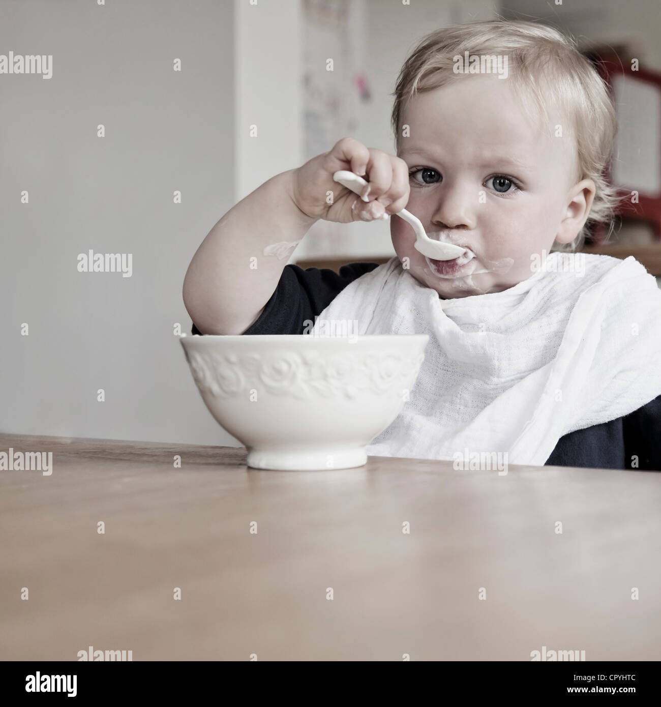 Junge (15 Monate) Essen Joghurt auf seinen selbst mit einem Löffel tragen Lätzchen Stockfoto