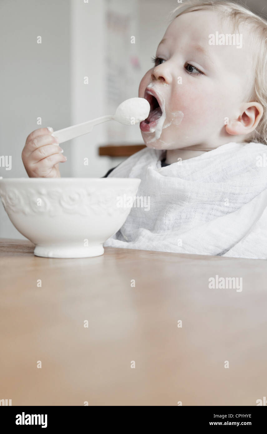 Junge (15 Monate) Essen Joghurt auf seinen selbst mit einem Löffel tragen Lätzchen Stockfoto