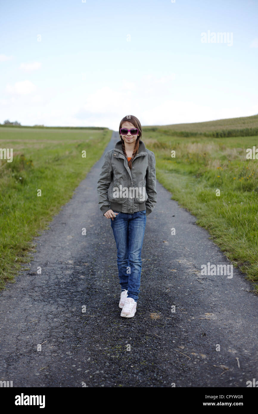 13 Jahre altes Mädchen in ein stilvolles Outfit mit rosa Sonnenbrille auf eine einsame Schotterstraße in ländlicher Umgebung Stockfoto