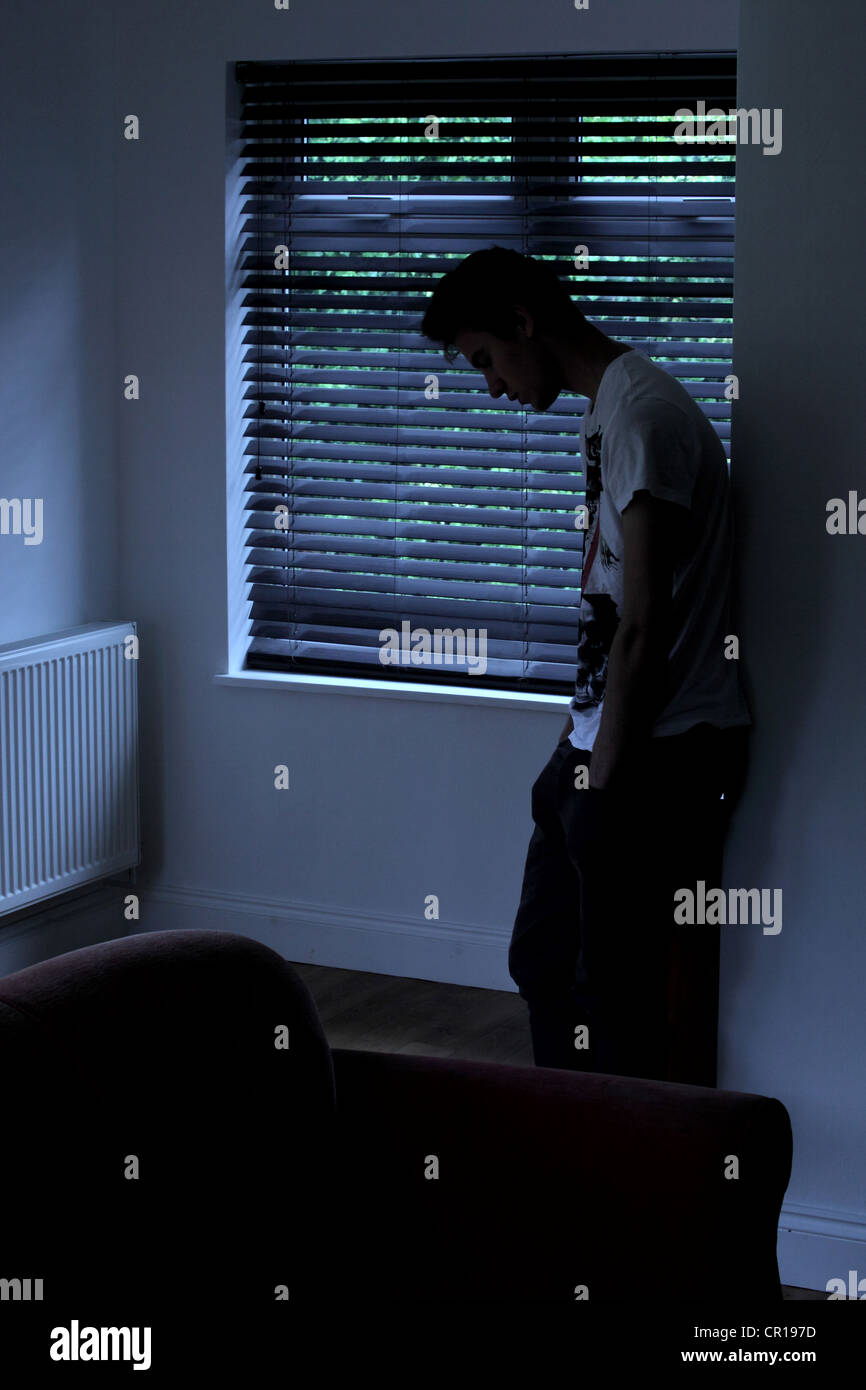 Junge Mann in einem dunklen Raum gegen die Wand hinter ihm schiefen ist ein Fenster blind. Modell und Eigentum (vom Fotografen) veröffentlicht. Stockfoto
