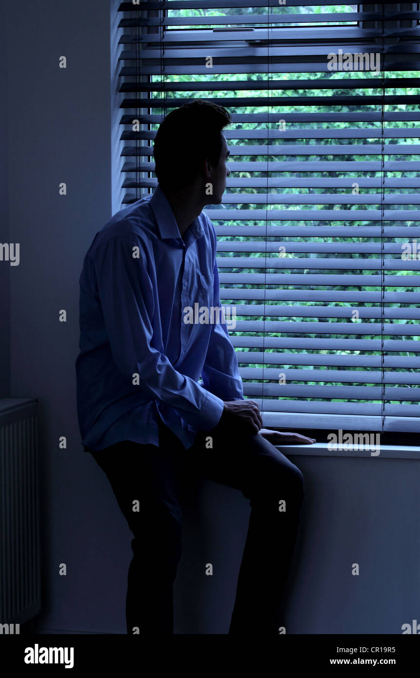 Junger Mann sitzt in einem dunklen Raum mit Blick durch ein Fenster Blind. Stockfoto