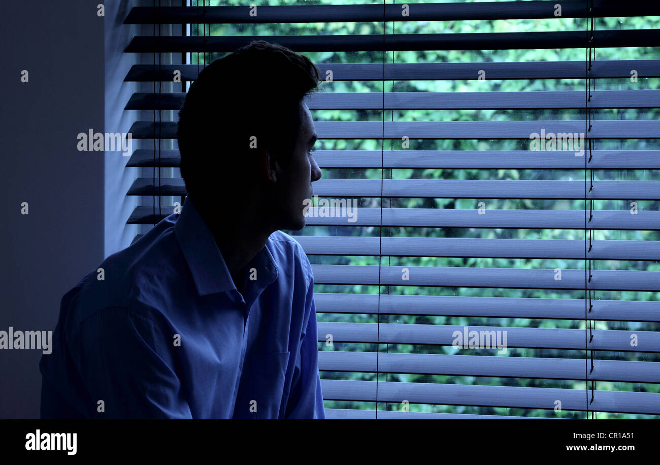 Junger Mann sitzt in einem dunklen Raum mit Blick durch ein Fenster Blind. Stockfoto