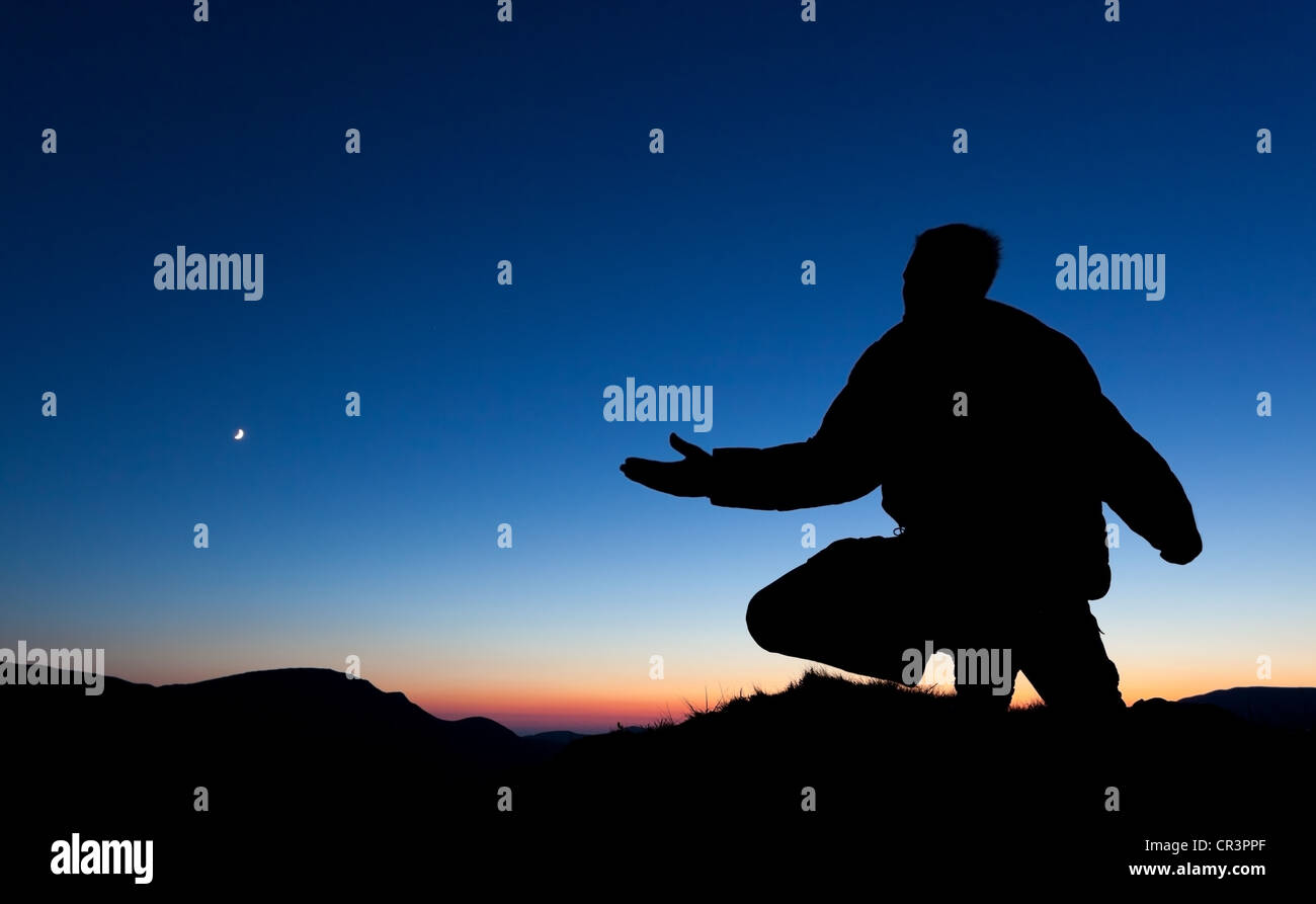 Mann, der auf dem Gipfel eines Berges bei Sonnenuntergang mit dem Mond am Himmel betet. Stockfoto