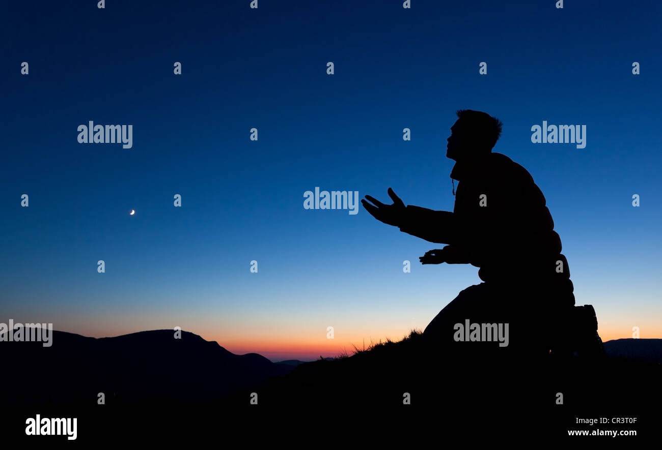 Man bittet Gott auf dem Gipfel eines Berges bei Sonnenuntergang mit dem Mond in den Himmel. Stockfoto