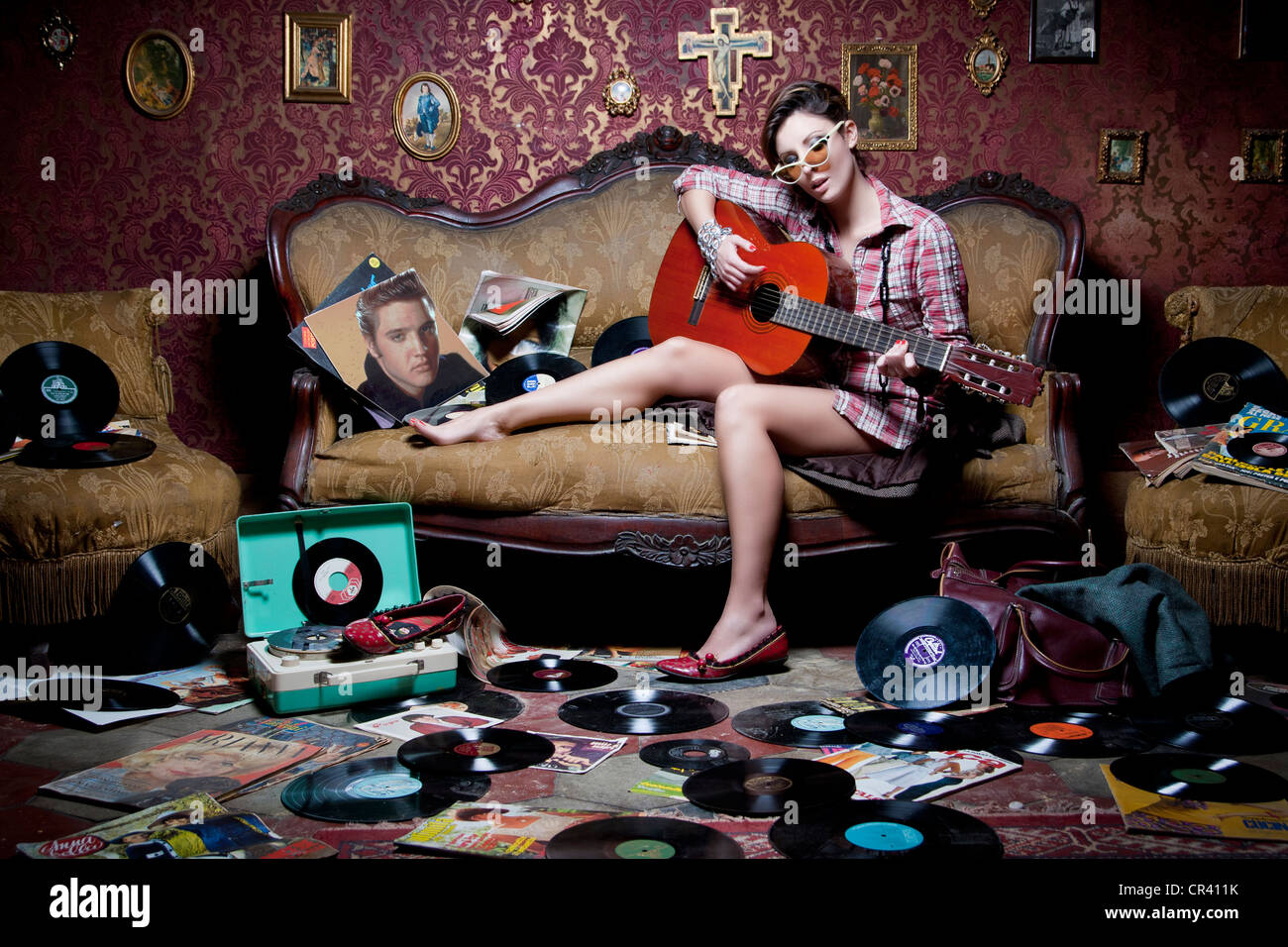 Mädchen spielt Gitarre auf ein altes Sofa mit vielen Vinyls und Zeitschriften auf dem Boden. Im Retro-Stil gedreht, perfekt für Veranstaltungen anzeigen. Stockfoto