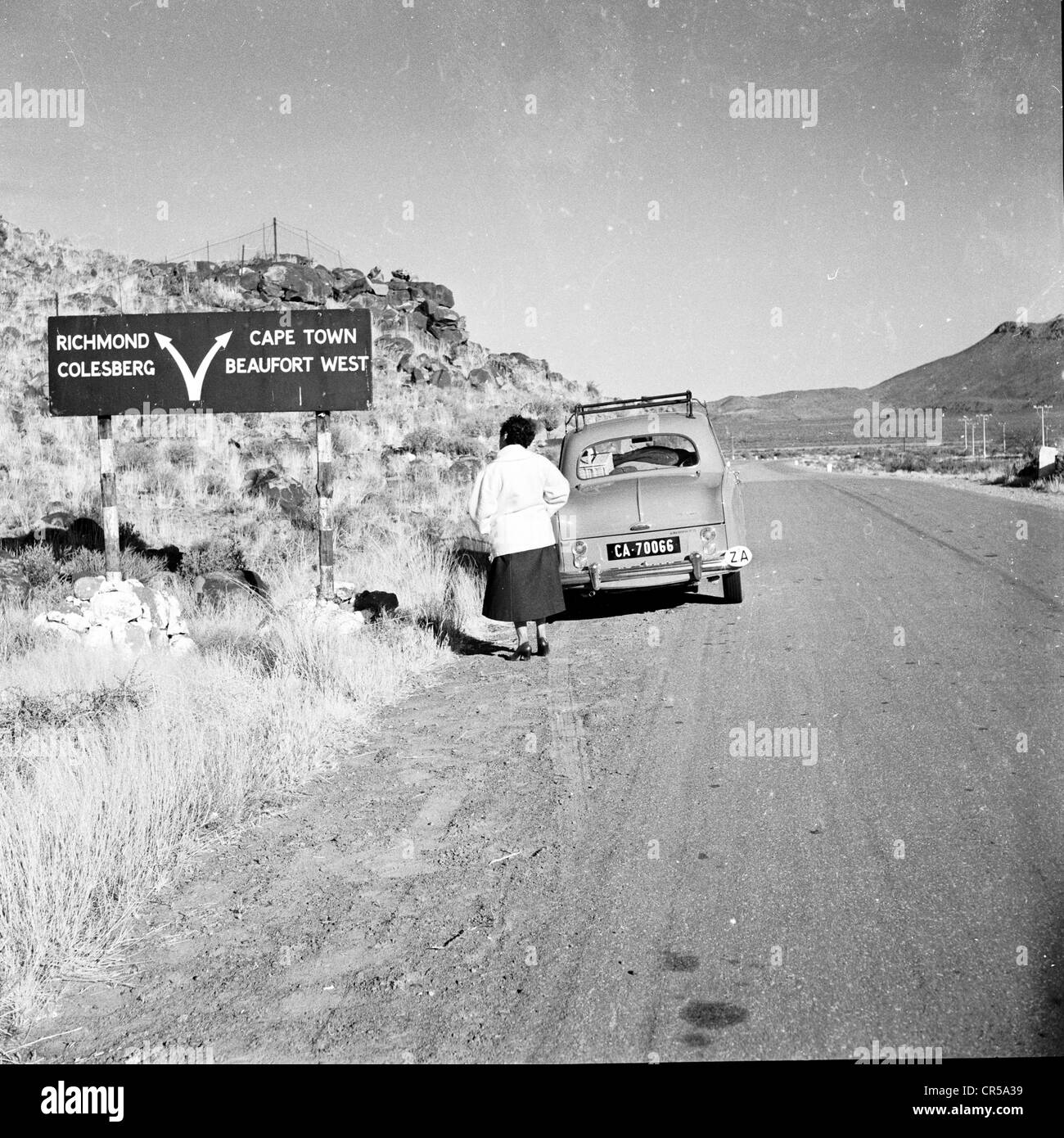 Südafrika, 50er Jahre. Eine Dame, die hinter ihrem kleinen Auto auf einer staubigen, leeren Straße im Outback des riesigen afrikanischen Landes stand, schaute auf eine Straße oder ein Richtungsschild und zeigte Pfeile zu verschiedenen Orten, darunter Kapstadt, Beaufort West, Richmond und Colesberg. Stockfoto