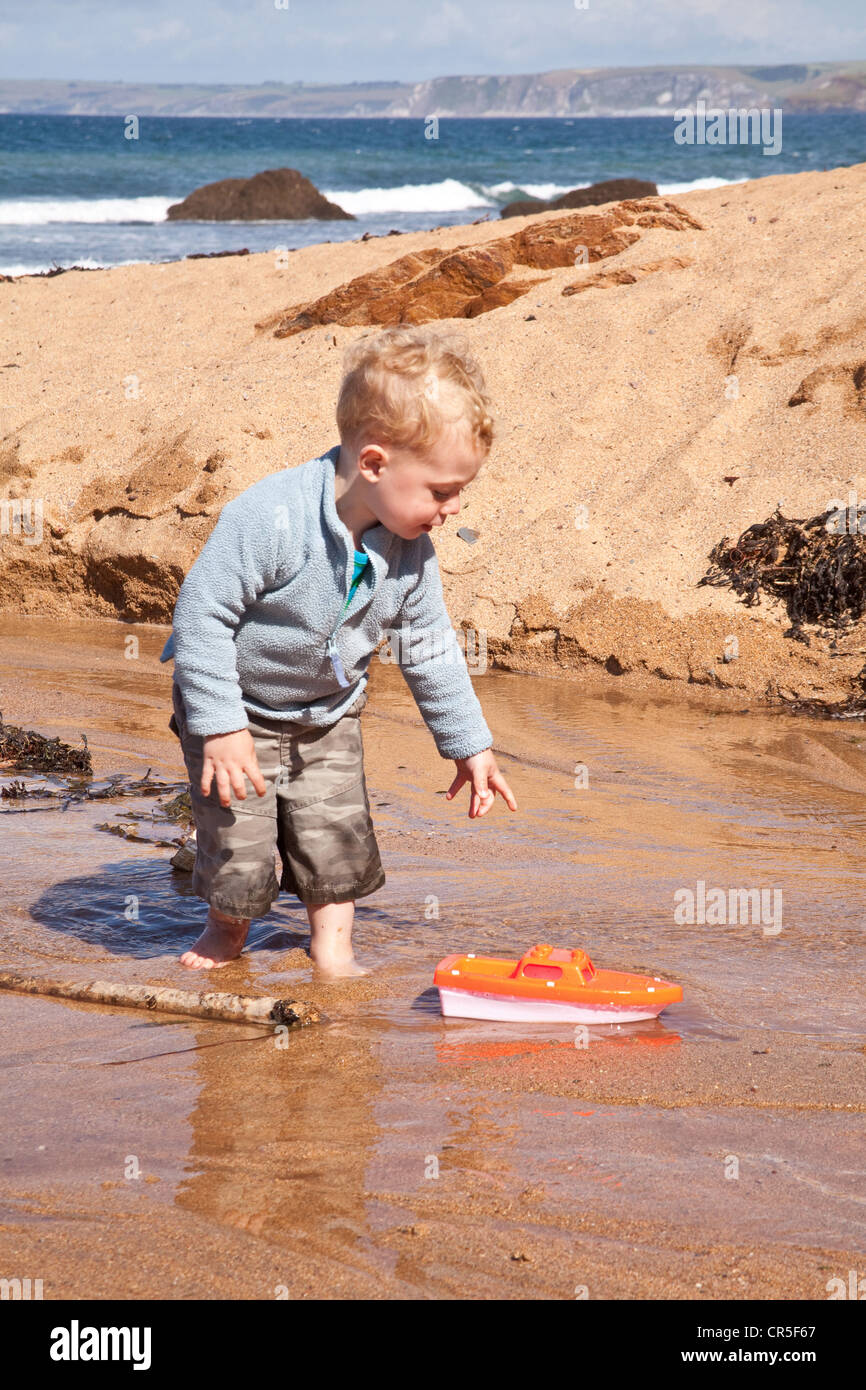 Kleiner Junge (zwei Jahre) spielen mit einem Spielzeugboot auf Hope Cove Beach, Devon, England, Vereinigtes Königreich. Stockfoto