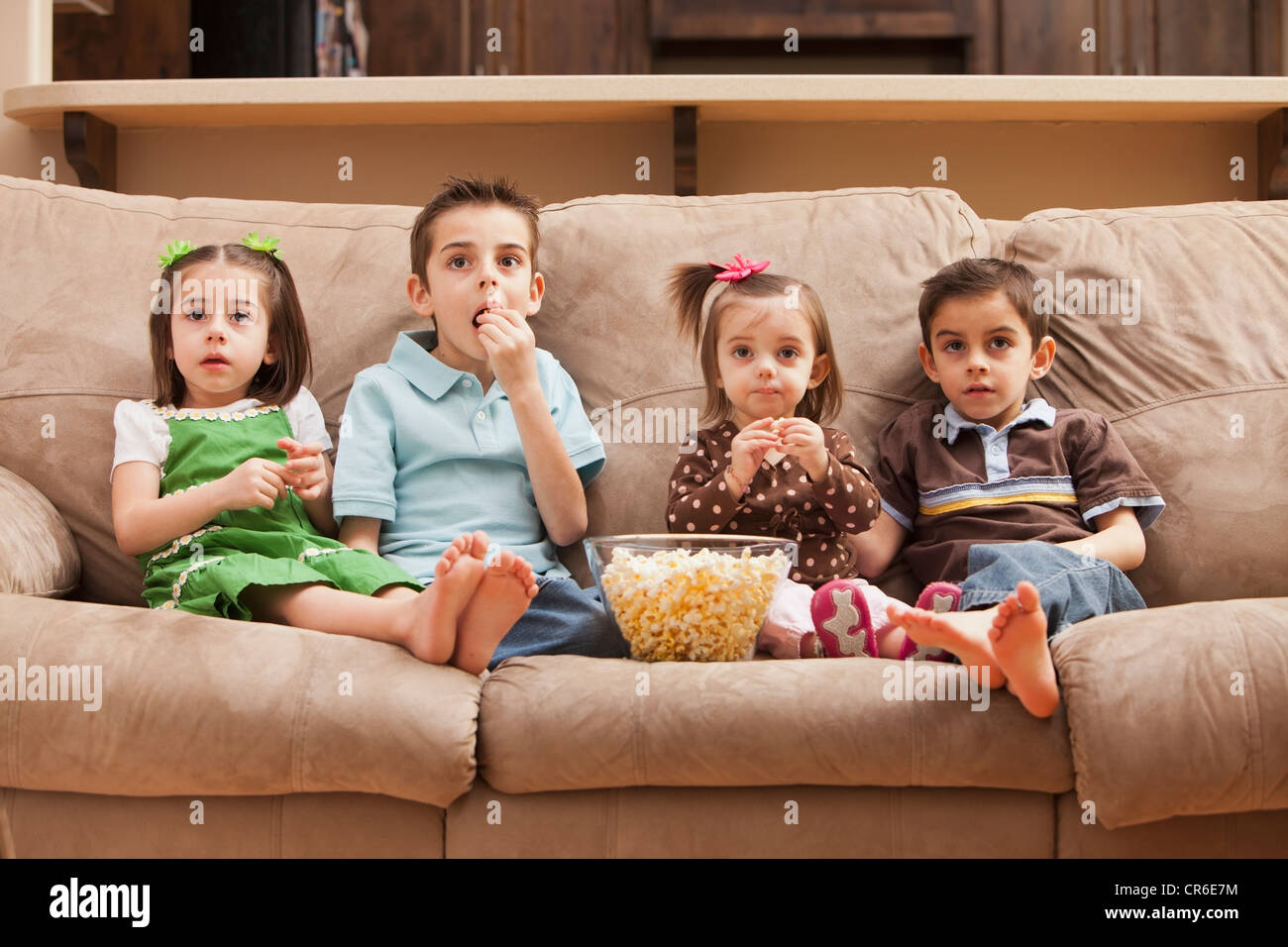 Frontansicht der Kinder (18-23 Monate, 4-5, 6-7, 8-9) sitzen auf der Couch vor dem Fernseher und Essen Popcorn Stockfoto