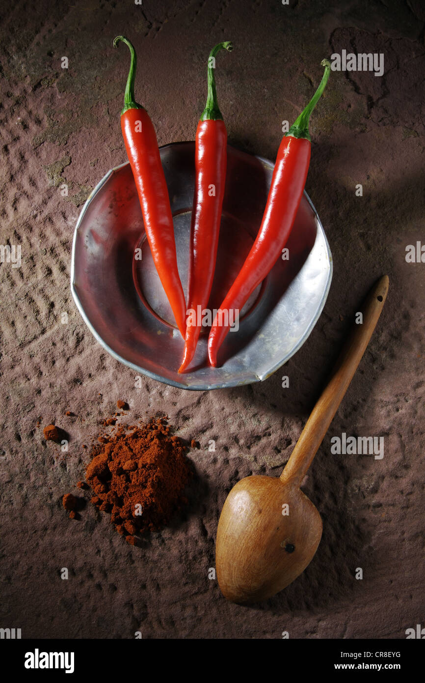 Drei Chili Paprika (Capsicum) auf einem Zinn-Teller mit einem Holzlöffel und Chili-Pulver auf einem rustikalen Steinoberfläche Stockfoto