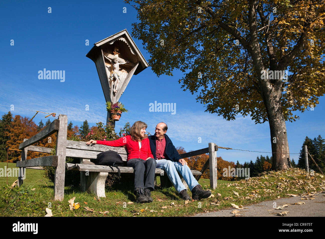 Paar, 45 und 55, ruht auf einer Bank vor einem am Wegesrand Kruzifix im Herbst, Alpine Vorberge, Bayern, Oberbayern Stockfoto