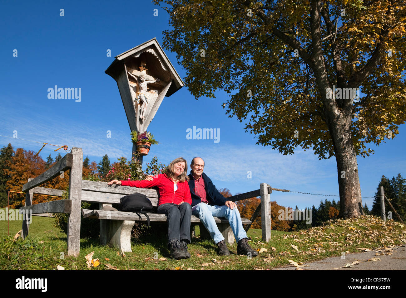 Paar, 45 und 55, ruht auf einer Bank vor einem am Wegesrand Kruzifix im Herbst, Alpine Vorberge, Bayern, Oberbayern Stockfoto
