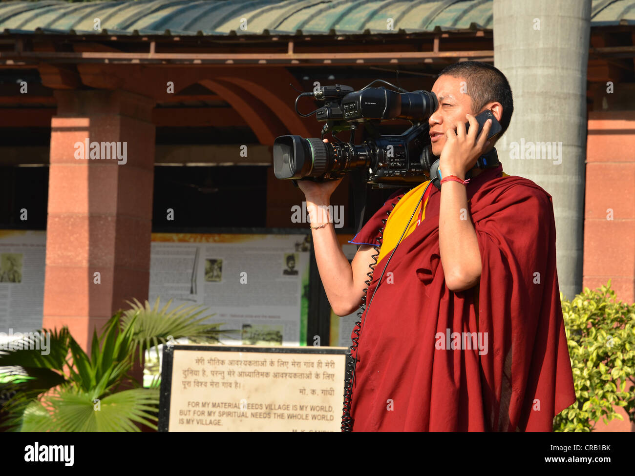 Tibetischer Mönch in ein rotes Gewand arbeitet als Kameramann mit einer großen Videokamera und einem Mobiltelefon, globale buddhistische Gemeinde Stockfoto