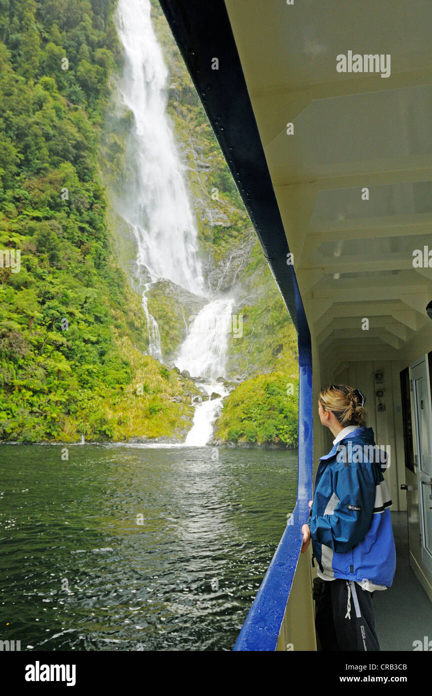 Frau lehnt sich an die Reling des Schiffes Fiordland Navigator, ein Wasserfall am Rücken, Doubtful Sound Kreuzfahrt, Fjord in der Stockfoto