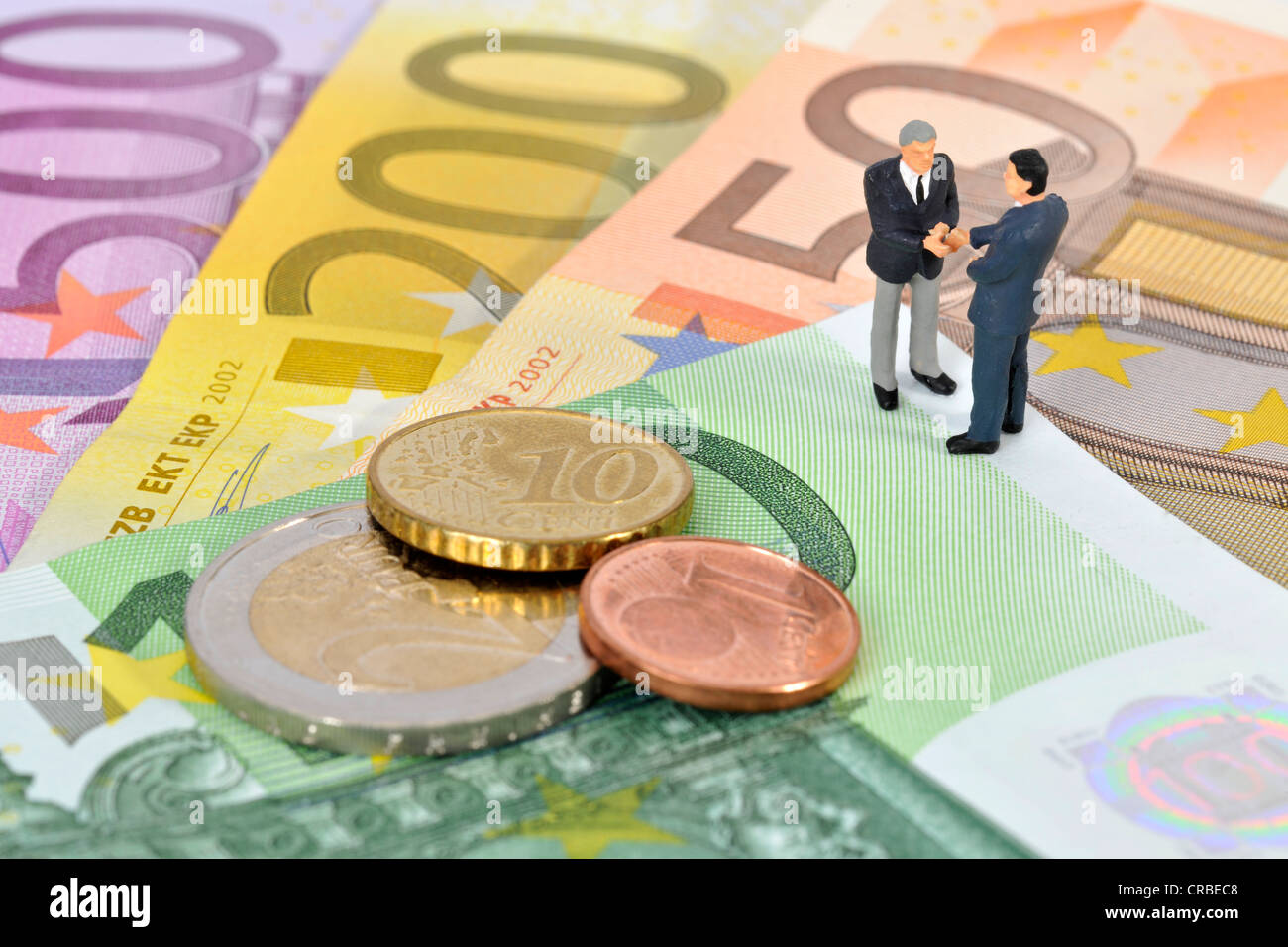 Miniatur-Figuren von Managern Händeschütteln beim stehen auf Euro-Banknoten und Münzen, symbolisches Bild für Unternehmen Stockfoto