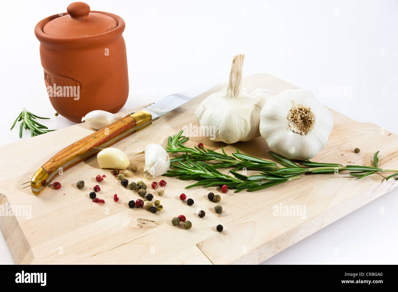 Knoblauch (Allium Sativum) auf einem Holzbrett mit Messer, Römertopf, Rosmarin und schwarzem und rotem Pfeffer Stockfoto