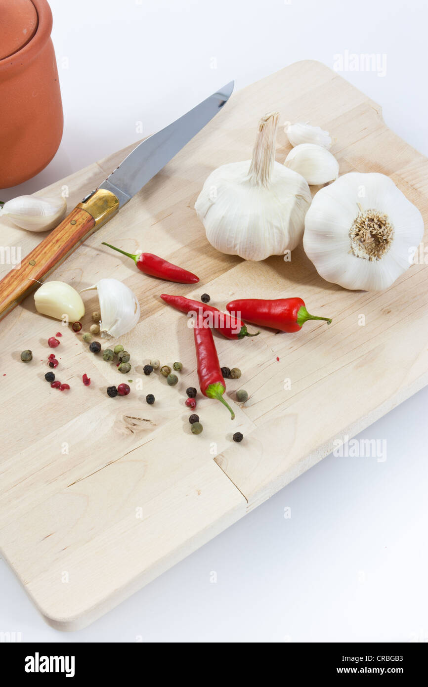Knoblauch (Allium Sativum) auf einem Holzbrett mit Messer, Tontopf, Chili und schwarzem und rotem Pfeffer Stockfoto